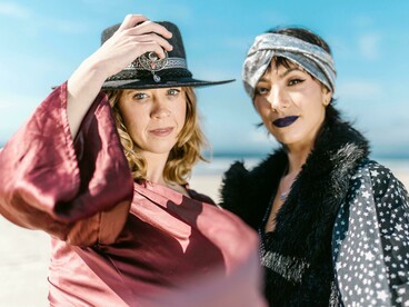 Duas mulheres sentadas na areia, com looks boho e acessórios marcantes, exploram a estética livre e criativa típica dos festivais onde estilo e personalidade caminham juntos na experiência