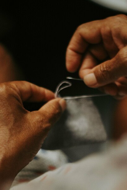 Mãos seguram agulha e linha enquanto costuram cuidadosamente um tecido. Remendar é um gesto simples que desafia a lógica do descarte. Muitas vezes, o que sustenta uma peça é invisível, tal como acontece com as reservas de paciência e coragem