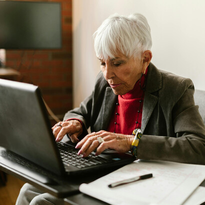 A senior woman in a gray blazer focused on her laptop while sipping coffee, reflecting demographic change and the active role of older generations in today’s digital society