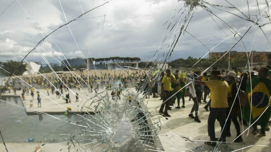 Por trás do vidro estilhaçado, manifestantes vestidos predominantemente de verde e amarelo ocupam a rampa de acesso ao Palácio do Planalto, durante os atos de 8 de janeiro de 2023. A destruição das sedes dos Três Poderes expôs a gravidade da ofensiva contra as instituições democráticas brasileiras perpetradas por estes mesmos representantes da extrema-direita