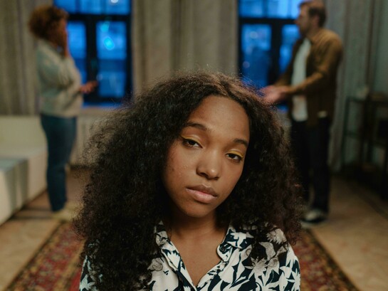 A concerned teenage girl with curly hair stands indoors while two adults argue in the blurred background
