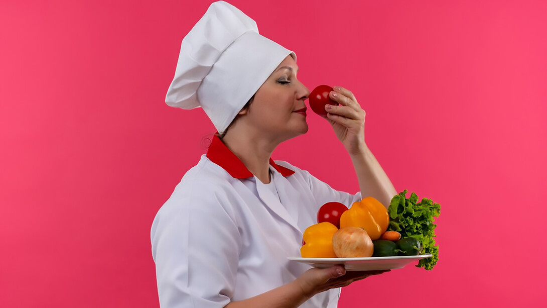 A middle-aged female chef in a chef's uniform, holding a plate of vegetables, sniffing a tomato in her hand