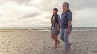 Pai e filha caminham lado a lado à beira-mar. Relações de afeto e cuidado também moldam a forma como homens envelhecem. Entre passos lentos e companhia constante, o cuidado aparece como vínculo e não apenas obrigação. Em muitos casos, são justamente as mulheres que sustentam redes emocionais e de cuidado ao longo da vida. O texto discute como masculinidade, afeto e cuidado influenciam até a expectativa de vida