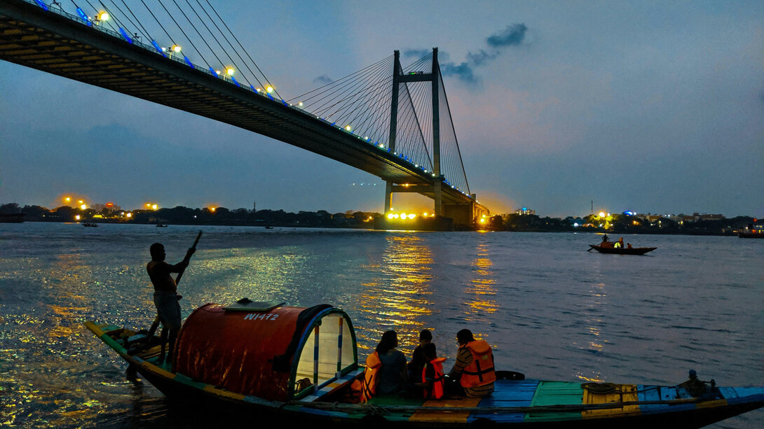 People riding boats on the water at night, with the vibrant city lights of Kolkata, the City of Joy, reflecting on the surface in West Bengal, India