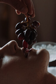Homem deitado em cima de uma mesa come uvas de olhos fechados 