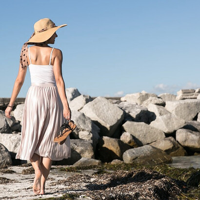 On the sandy shores, a woman radiates poise, dressed in a white skirt and holding black leather flat sandals, embracing the coastal scenery with a harmonious blend of elegance and relaxation