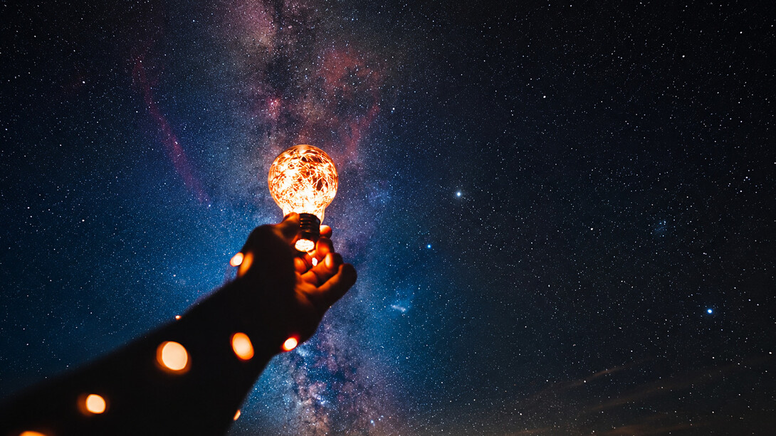 Uma mão ergue uma lâmpada acesa diante de um céu estrelado, em uma metáfora para o surgimento de novas ideias e descobertas. A inovação científica nasce da curiosidade, mas só transforma a sociedade quando o conhecimento é desenvolvido, protegido e colocado em prática. A mão segurando uma lâmpada acesa diante de um céu estrelado remete à criatividade, ao conhecimento e ao potencial transformador da ciência. Em vez de representar o ambiente laboratorial, ela sintetiza visualmente a ideia de que uma descoberta pode iluminar novos caminhos quando é convertida em inovação