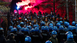 A heated standoff unfolds between riot police and protesters, the air thick with the anticipation of conflict and the weight of personal struggles behind every helmet