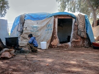 Tenda in un campo profughi e un bambino inginocchiato davanti a una stufa