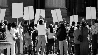 A crowd of protesters gathers at a political demonstration, holding signs and expressing their frustration
