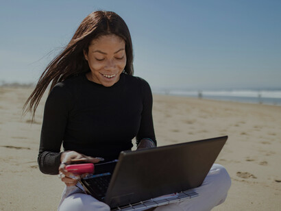 A woman in a black long-sleeve shirt works on her laptop at a beachfront, embracing the digital nomad lifestyle