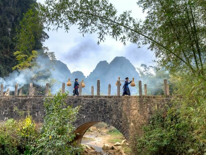 Two villagers crossing a weathered bridge with towering mountains in the background, reflecting the strong spirit of communities shaped by hardship and hope
