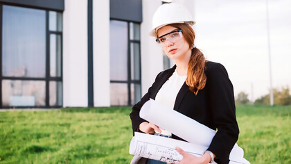 A female architect stands confidently in front of a building, holding architectural plans in her hands