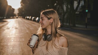 A smiling blonde woman enjoys a warm drink while listening to music or language lessons through her headphones