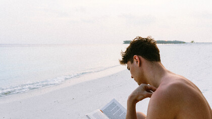 Rapaz lê livro em beira da praia de frente para o mar