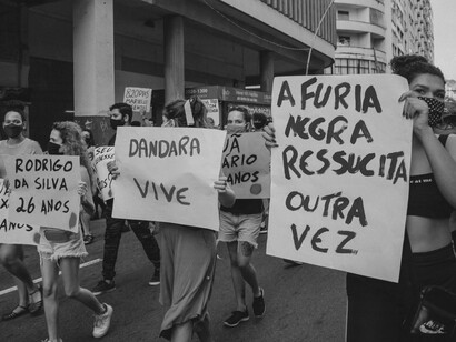 Manifestantes carregam cartazes com frases como "A fúria negra ressuscita outra vez" (essa é uma das frases mais emblemáticas do rap brasileiro; ela faz parte da música "Capítulo 4, Versículo 3", lançada em 1997 pelos Racionais MC's no clássico álbum "Sobrevivendo no Inferno"); "Dandara vive" (Dandara dos Palmares foi uma grande liderança feminina e guerreira do Quilombo dos Palmares, no Brasil colonial. Companheira de Zumbi dos Palmares, ela dominava técnicas de capoeira, lutava na linha de frente contra as tropas portuguesas e ajudava a criar estratégias de resistência); e "Rodrigo da Silva 26 anos" (homem negro, trabalhava como garçom, foi assassinado pela Polícia Militar do Rio de Janeiro enquanto esperava sua esposa e filhos retornarem para casa, em um dia chuvoso, quando ele os aguardava com um guarda-chuva que foi confundido com um fuzil por policiais que atiraram contra ele, conforme relato de testemunhas). O artigo destaca a importância das mobilizações sociais contra diferentes formas de exclusão e violência