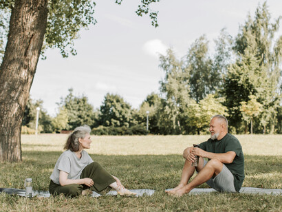 A gray-haired man and a woman sitting barefoot on mats, in a park