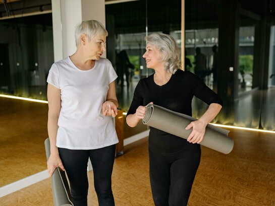 Two senior women chatting and smiling while holding yoga mats inside a modern fitness studio
