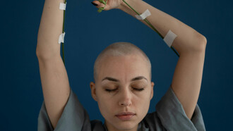 A shaved young woman with eyes closed and arms reaching up