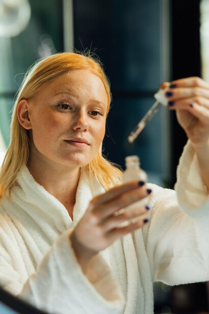 A woman in a white robe holding a dropper in front of a mirror, representing the gap between promising ingredients and what the skin can actually absorb