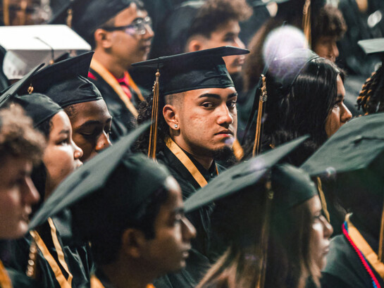 Un joven apelando  a la cámara con mirada seria en el día de su graduación, junto al resto de sus compañeros