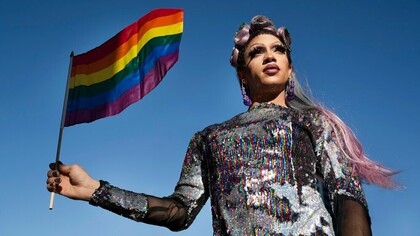 A man in drag wearing a sparkly outfit and waving the rainbow flag