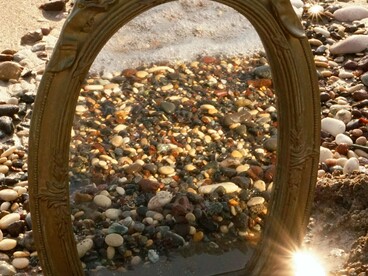 Um espelho de moldura ornamentada repousa entre as pedras à beira-mar, refletindo a paisagem ao redor. Na simbologia de Oxum, o espelho vai além da beleza exterior: convida ao encontro com a própria essência e à descoberta daquilo que permanece em nós, mesmo diante das perdas