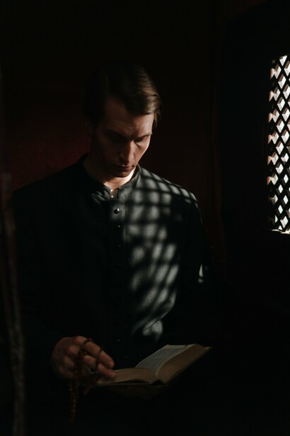 A priest reflecting in the stillness of a confessional while reading from a sacred text, symbolising the importance of restoring ministry above material interests