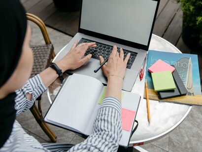 Pessoa trabalha diante de um laptop colocado sobre a mesa. Entre a escrita e as ferramentas digitais, permanece o elemento que a tecnologia não pode reproduzir: a experiência de quem cria