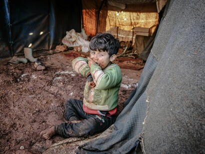 A child huddles in the dirt with little more than a torn sweater for warmth—revealing the devastating human cost of aid that never delivers on its promises