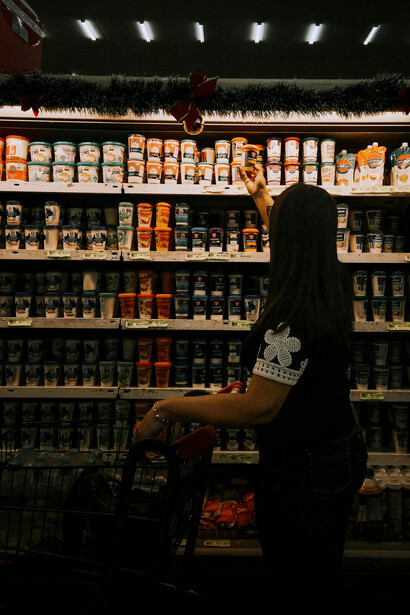 A woman with a shopping cart walks through a dimly lit grocery aisle, silhouetted against shelves filled with neatly arranged products