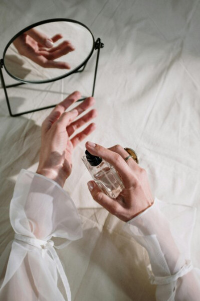 A woman applies perfume to her wrist while resting her arm on white sheets