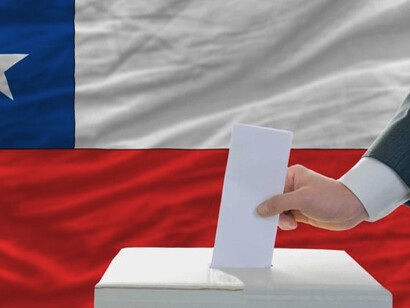 Chile celebró recientemente la primera vuelta de sus elecciones generales el 16 de noviembre de 2025, para elegir un nuevo presidente, todos los miembros de la Cámara de Diputados y la mitad de los escaños del Senado. Una mano depositando su voto en una urna, con la bandera nacional de Chile de fondo