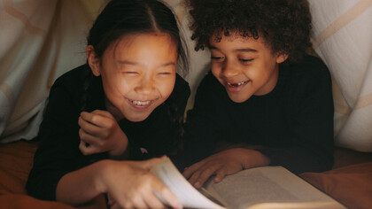 Crianças dentro de cabana feita com lençois lendo livro engraçado