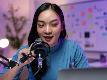 A young woman records a podcast in her home studio, speaking into a microphone while monitoring content on her laptop, with a whiteboard visible in the background