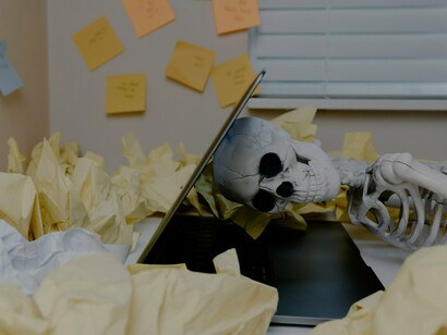A skeletal form slumped over its work, lost in a sea of notes and unfinished projects; the ultimate warning of burnout, when the worker is left with nothing but remnants of their former self
