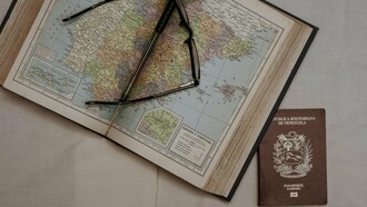 A pair of glasses rests on an open map, alongside a passport from the Bolivarian Republic of Venezuela, visually capturing the forced journeys and global scattering of Venezuelan exiles as they navigate new lives far from their homeland
