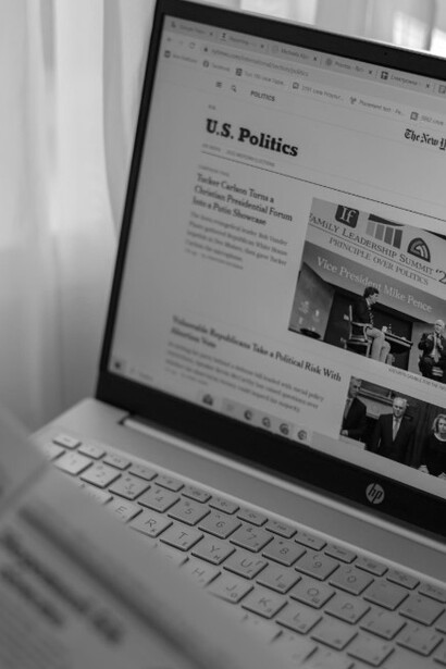 A laptop displaying news sits on a desk beside a man holding and reading a newspaper, both filled with headlines about American politicians