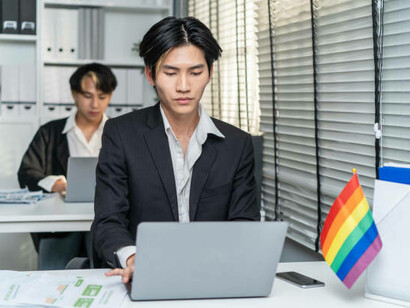 Asian gay businessmen working together in an office, exuding happiness and confidence. The attractive male LGBTQ+ colleagues, dressed in formal attire, sit at the table, using laptops while proudly embracing their identity at the workplace