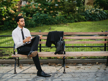 A man in a suit and white long-sleeve shirt sits on a brown wooden bench, searching for a job