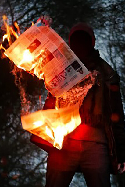 A person holding a burning newspaper symbolizes the crisis of democracy in the face of media manipulation, information pollution, and the ongoing struggle to build democratic resilience