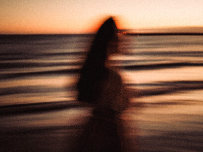 Retrato difuminado de perfil de una persona con cabello largo frente al mar durante el atardecer, con tonos cálidos y efecto de movimiento