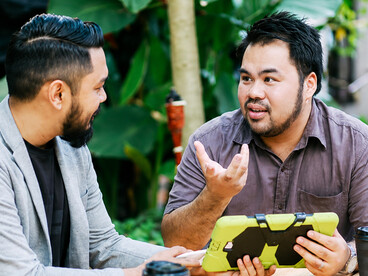Over coffee in a plant-filled café, two men engage in a thoughtful conversation about the challenges they are facing