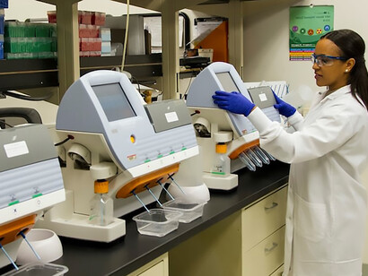 In the Cancer Genomics Research Laboratory, part of the National Cancer Institute's Division of Cancer Epidemiology and Genetics (DCEG), a technician adeptly operates a series of desktop genomic sequencing machines, underscoring the meticulous process of DNA genotyping and sequencing. A woman stands confidently at the forefront of this cutting-edge technology, embodying the intersection of expertise and innovation in genetic research