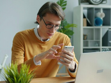 Um homem usa o celular enquanto permanece sentado à mesa, cercado por objetos de trabalho. A tela concentra sua atenção e traduz a presença constante da tecnologia no cotidiano, um dos fatores que o artigo associa ao distanciamento da experiência emocional. Em meio à notificações, tarefas e estímulos permanentes, a hiperconectividade pode aproximar o indivíduo do mundo digital e, paradoxalmente, afastá-lo da própria experiência