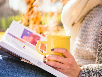 Donna con una tazza in mano mentre legge un libro all'aperto: "Se quella spinta a scappare da te stesso, fosse un movimento verso le parti di te che non conosci?"