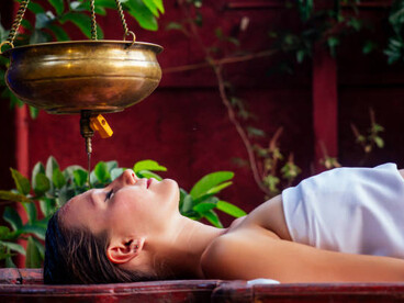 Shirodhara oil therapy, an alternative healing treatment, with a beautiful Caucasian woman receiving the therapy while lying on a wooden table in an Indian wellness salon