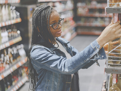 A young woman stands in a supermarket aisle, overwhelmed by the endless food options as she tries to decide what to buy