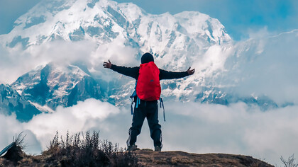 A man stands with both hands raised on a mountain cliff, overlooking the snow-covered peaks of the Annapurna range in Nepal