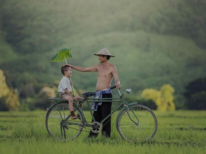 An Asian man is standing near his bicycle in the middle of the field and giving his child a natural but also comical head shield, it's the type of parent children appreciate